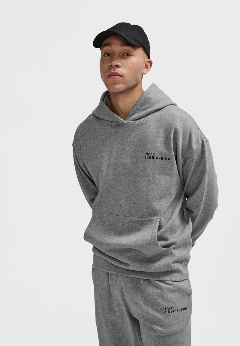 Młody mężczyzna w szarej bluzie z kapturem i pasujących dresach z czarnym napisem "HALO 196819721981", oraz czarnej czapce, stojący na białym tle.