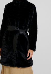 Sort faux fur frakke med høj krave, ribbet tekstur og et bredt, fladt sort bælte bundet om taljen, med en blød, plysset overflade.