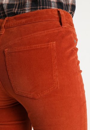 Gros plan sur un pantalon en velours côtelé orange brûlé avec une poche arrière, porté avec une chemise écossaise noire, blanche et marron rentrée dedans.