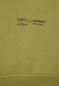 Tela verde oliva con texto impreso en negro que incluye "23ENGINEERED", "R/DASSEMBLY", información de versión y coordenadas sobre una costura cosida.