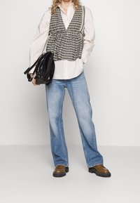 Chaleco de cuadros tipo "houndstooth" sobre una camisa blanca, combinado con unos jeans azules holgados y unos zapatos gruesos marrones, sosteniendo un bolso negro de charol.