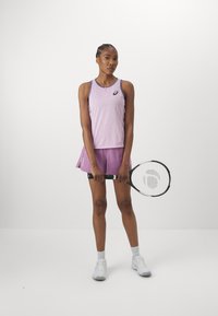 Lilla trenings tank-top med et teksturert mønster, kombinert med en matchende plissert skjørt. Modellen holder et tennisracket.