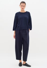 Tengerészkék kötött pulóver széles ujjakkal, kerek nyakkivágással és bordás aljjal, laza tengerészkék nadrággal és fekete slip-on cipőkkel párosítva.