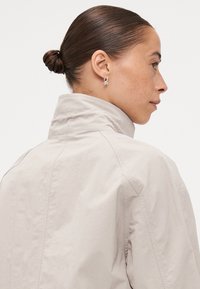 Light grey, high-collared jacket made from smooth fabric, featuring visible seams and a relaxed fit, with a minimalistic design and no closures.