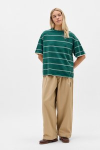 Grünes gestreiftes T-Shirt mit weißen horizontalen Linien, lässige Passform, kombiniert mit beigefarbenen weit geschnittenen Hosen. Lässiges Schuhwerk rundet das Outfit ab.