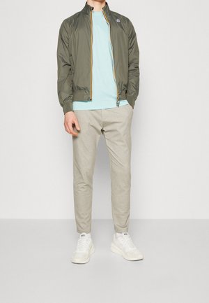 Hombre con chaqueta con cremallera color verde oliva, camiseta azul claro, pantalones beige y zapatillas blancas con calcetines blancos sobre un fondo liso.