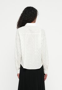 Biała bluzka z długim rękawem z subtelnym wzorem kropek w kolorze złotym. Posiada szpiczasty kołnierzyk oraz lekko zaokrąglony tył.