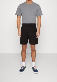 Šedé bavlněné tričko v kombinaci s tmavými bavlněnými šortkami. Model má na sobě bílé ponožky a barevné tenisky s texturovanými materiály a tkaničkami.