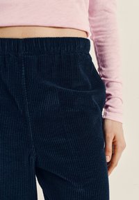 Žmogus, dėvintis tamsiai mėlynas, aukštu liemeniu pasiūtas korduro kelnės ir šviesiai rožinę ilgomis rankovėmis marškinių, dešinė ranka atsipalaidavusi šalia kūno.