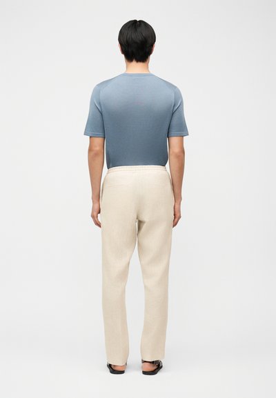 Άνδρας με ανοιχτό μπλε κοντομάνικο πουκάμισο, μπεζ παντελόνι και μαύρα σανδάλια, στέκεται με την πλάτη προς το μέρος μας σε απλό λευκό φόντο.