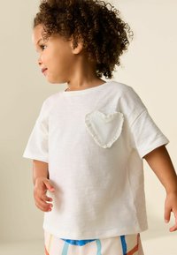 Weißes T-Shirt mit kurzen Ärmeln aus weichem Stoff, mit einem strukturierten, herzförmigen Aufnäher mit Rüschensaum auf der linken Brust.