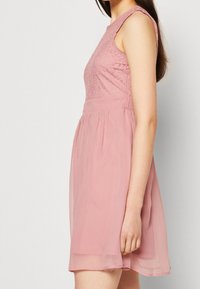 Robe sans manches rose avec un corsage en dentelle texturée, jupe en chiffon fluide et taille froncée. Couleur douce et tissu léger et aérien.