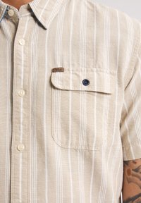 Camisa beige de manga corta con rayas verticales blancas, cierre frontal de botones y un bolsillo en el pecho con un botón azul marino y etiqueta de la marca.
