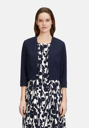 Veste cropped marine à manches trois quarts, en tissu lisse, au design à l'avant ouvert. Portée sur une robe blanche ornée d'un motif floral noir.