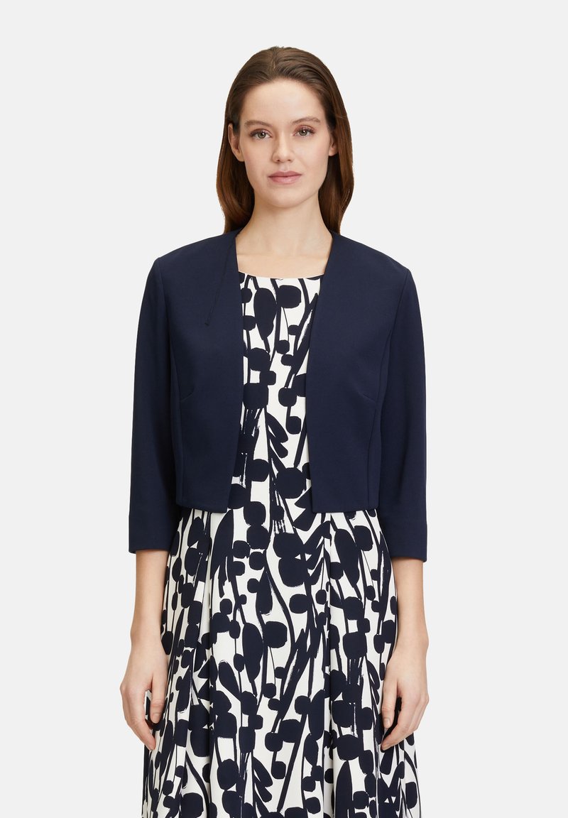Veste cropped marine à manches trois quarts, en tissu lisse, au design à l'avant ouvert. Portée sur une robe blanche ornée d'un motif floral noir.