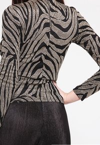 Czarna bluzka z długim rękawem w metaliczne złote paski w zebrę, charakteryzująca się dopasowanym krojem i zapięciem z tyłu na zamek błyskawiczny. Połączona z czarnymi leginsami w prążki.