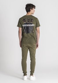 Camiseta verde oliva con un gráfico de tres perros Doberman y el texto "Vavanagh". Pantalones ajustados a juego y zapatillas blancas.
