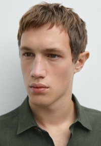 Close-up of a young man with short brown hair and light skin, wearing a dark green collared shirt. Features include smooth skin and neutral expression.