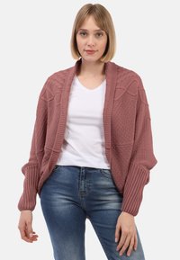 Cardigan tricoté en rose poussière avec des motifs texturés, devant ouvert et poignets côtelés, superposé à un T-shirt blanc et un jean bleu.