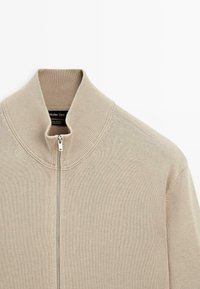 Beige Reißverschluss-Pullover mit hohem Kragen, aus 100% Baumwolle gefertigt. Verfügt über feine Rippungen und eine subtile Textur, mit einem silbernen Reißverschluss-Detail.