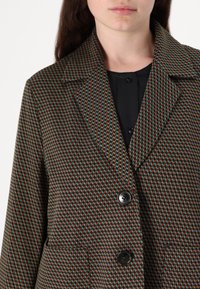 Mønstret blazer med et geometrisk design i rødt, grønt og sort. Den har en klappet krave og to knapper foran.