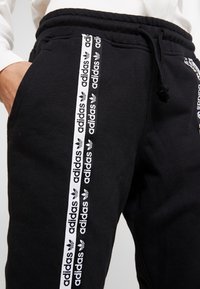 Svarta träningsbyxor med en texturerad yta har vita sidorekler med "adidas"-logotypen och trefoil-mönstret, samt ett elastiskt midjeband.