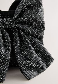 Juoda audinio kaspino su tekstūruota paviršiaus, papuošta daugybe mažų krištolo akutės, kurios sukuria blizgantį efektą visame medžiagoje.