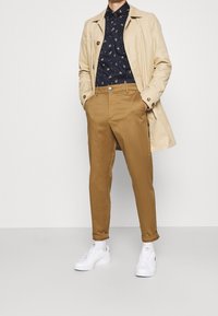 Trench coat beige sopra una camicia a fantasia blu navy, abbinato a pantaloni marroni e sneakers bianche. Caratteristiche includono bottoni e polsini arrotolati.