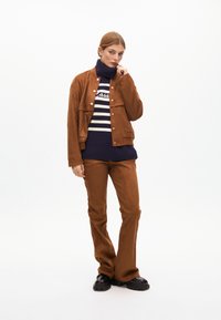 Hnědá semišová bunda s cvoky, námořnický pruhovaný rolák, hnědé přiléhavé kalhoty a černé platformové boty. Minimalistické pozadí.