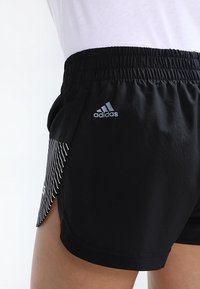 Svarta träningsshorts med elastisk midja, som har en silver Adidas-logotyp och vita vertikala ränder på sidan.