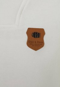 White cotton fabric with a brown leather patch featuring embossed text "CBJ" and "Cipo & Baxx Denim Collection." Simple, textured design.
