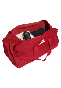 Bolsa de lona roja hecha de un tejido resistente. Cuenta con múltiples asas, cierre de cremallera y compartimentos interiores. Contiene una botella de agua negra y ropa.