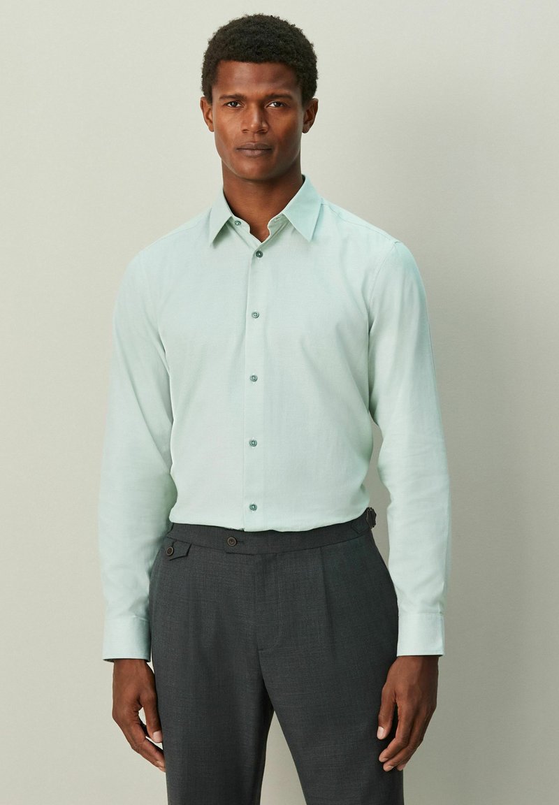 Moški, oblečen v svetlo zeleno srajco z dolgimi rokavi, zašito v temno sive po meri prilagojene hlače, na enobarvnem ozadju.