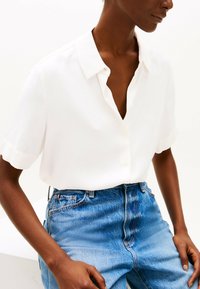 Blusa bianca a maniche corte con colletto, dal taglio rilassato e una sottile lucentezza, abbinata a jeans in denim blu a vita alta.