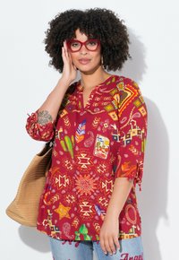Blusa roja con estampado que presenta diseños geométricos e inspirados en la naturaleza, con escote en V y mangas fruncidas. La modelo sostiene un bolso de tejido.