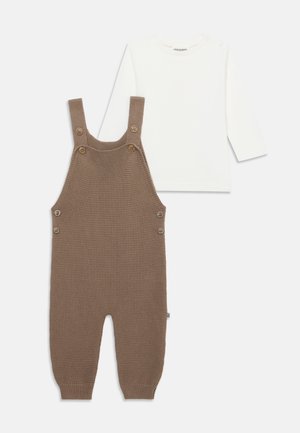 Salopette bébé marron en maille, avec des boutons aux bretelles et sur les côtés, associée à une chemise blanche à manches longues sur un fond uni.