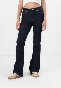 Jeans de mezclilla azul oscuro acampanados con costuras doradas, cinco bolsillos y cierre de botón. La tela es gruesa y texturizada.