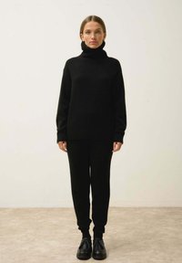 Musta neuleet turtleneck-pusero ja yhtenevät tiukat housut, valmistettu pehmeästä materiaalista, yhdistettynä mustiin nauhallisiin kenkiin. Yksinkertainen tausta.