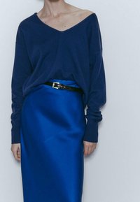 Laivastonsininen villapaita, jossa on olkapäät paljastava V-pääntie, yhdistettynä kiiltävään siniseen satiinihameeseen ja mustaan vyöhön, jossa on suorakaiteenmuotoinen solki.