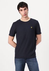 Tricou bleu marin cu mânecă scurtă, cu guler bej și un mic logo galben pe piept, realizat dintr-un material moale și neted.