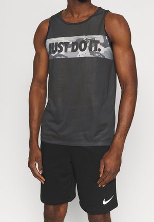 Mand iført sort ærmeløs skjorte med teksten "JUST DO IT." og sorte shorts med hvid Nike-logo, stående mod en ensfarvet baggrund.