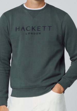 Mężczyzna w ciemnozielonym swetrze Hackett London z ręką w kieszeni, w połączeniu z białymi spodniami, na jednolitym tle.