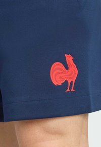 Granatowe szorty o teksturowanym wykończeniu, z wyraźnym czerwonym logo koguta na dolnej stronie. Materiał jest gładki i lekki.