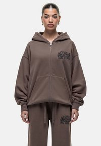 Felpa con zip marrone, dotata di ampie tasche frontali e logo stampato sul petto. Il tessuto è morbido, con una vestibilità rilassata, cappuccio e spalle scese.