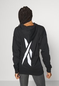 Czarna bluza z kapturem z dużym białym logo na plecach. Posiada luźny krój i ściągacze przy rękawach, wykonana z miękkiego, teksturowanego materiału.