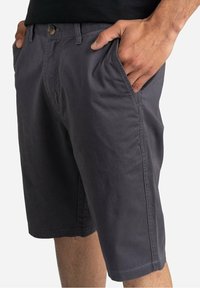 Pantaloni corti in cotone grigio con vestibilità rilassata, tasche laterali e chiusura con bottone. Il tessuto ha una consistenza liscia e un aspetto leggermente sgualcito.