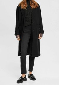 Abrigo de lana negro de corte holgado, con cierre de un solo botón y dos bolsillos frontales. Combinado con un suéter de punto verde oscuro y jeans negros.