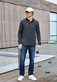 Schwarzes Langarm-Polo mit strukturiertem Muster und schwarzem Kragen, kombiniert mit blauen Jeans, weißen Sneakern und einer hellbeigen Kappe.