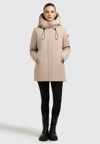 Giaccone beige imbottito con cappuccio, cerniera frontale, tasche laterali e coulisse regolabile. Indossato con leggings neri e stivali neri.