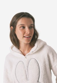 Biała rozpinana bluza z kapturem wykonana z miękkiego, puszystego materiału, z przesadnym szarym konturem twarzy królika z przodu.